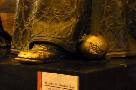 Fuß einer Bronzestatue, daneben ein Granatapfel aus Bronze, in dem eine Schwertspitze steckt.