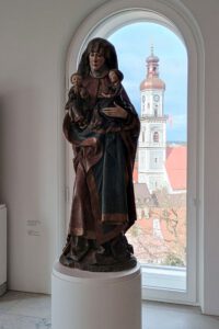 Madonnenstatue im Diözesanmuseum Freising, im Hintergrund ist durch das Fenster ein Kirchturm zu sehen.