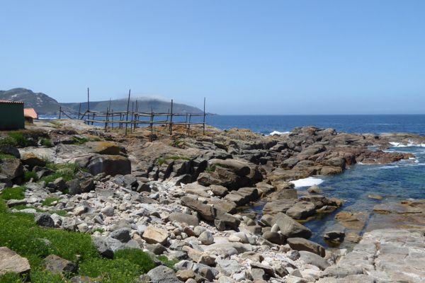 Hölzernes Trockengestell für Fische am Ortsrand von Muxia, Galicien, mit Blick auf Meer und Berg.